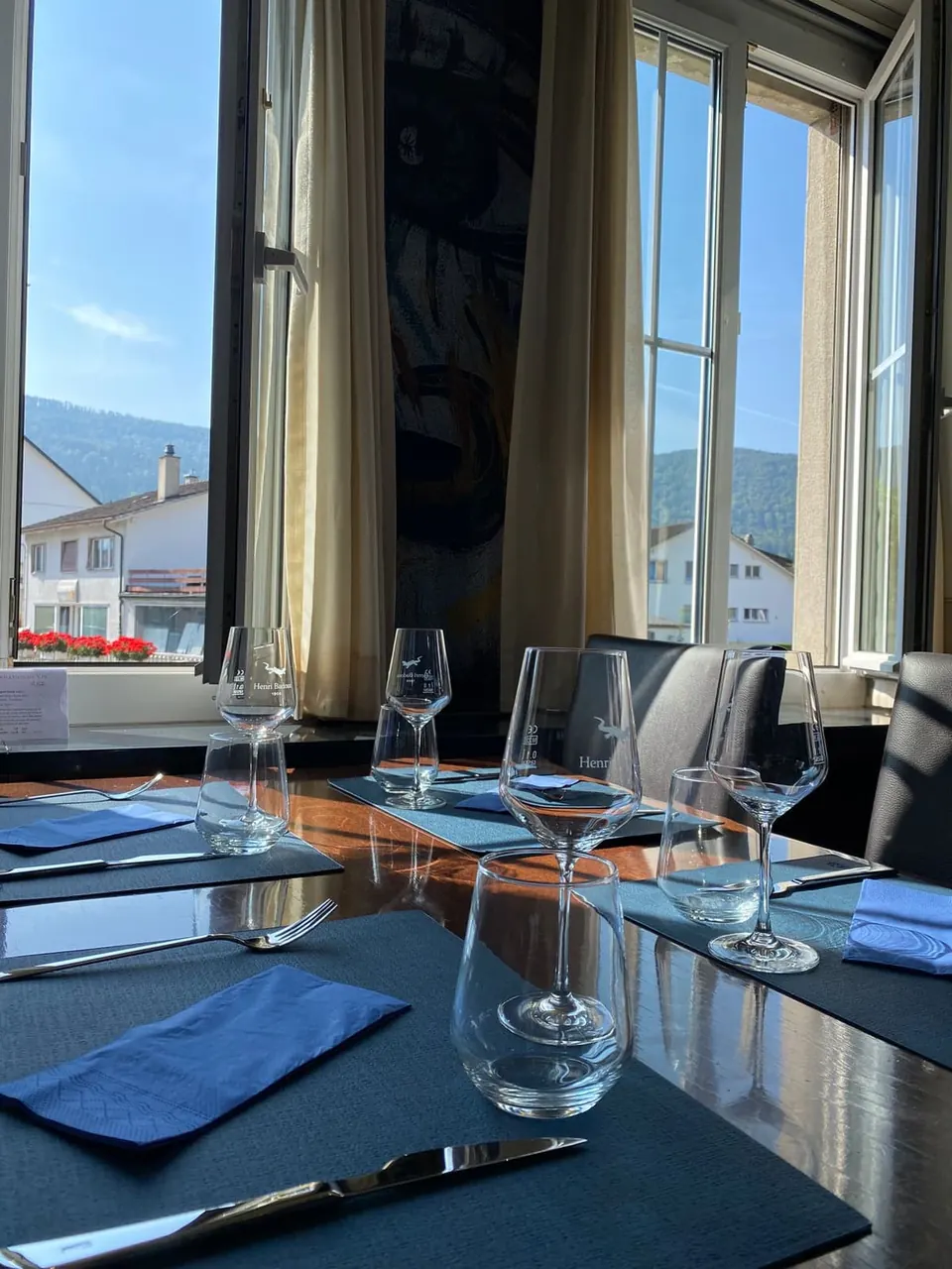 Une table dressée près de la fenêtre : verres à pied, sets et serviettes bleu nuit, les collines au loin.